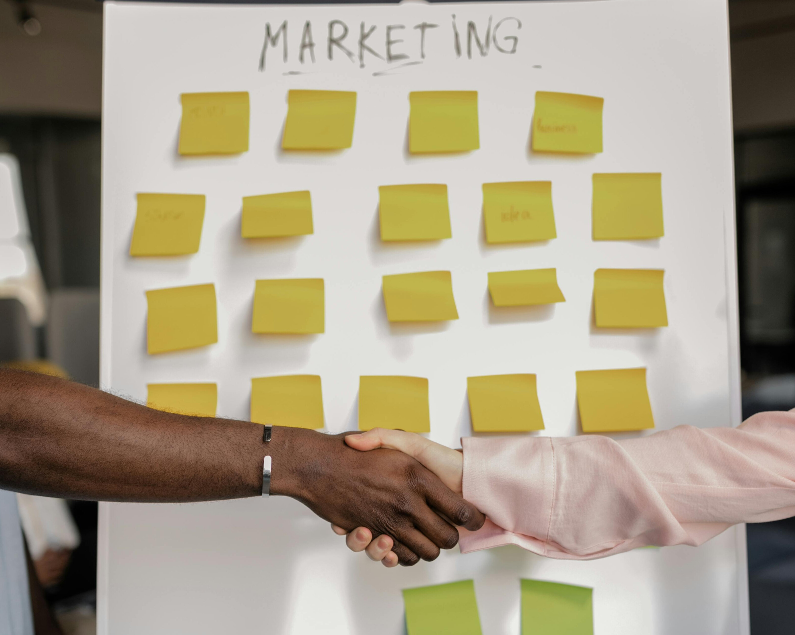 Two individuals shaking hands in front of a whiteboard covered with yellow sticky notes labeled “MARKETING,” representing collaboration and opportunity in learning digital marketing.